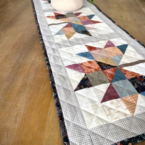 May include: A rectangular quilted table runner featuring a repeating star pattern made of various fabric patches in shades of blue, orange, red, and cream. The runner has a gray and white checkered border and a dark floral print edge.