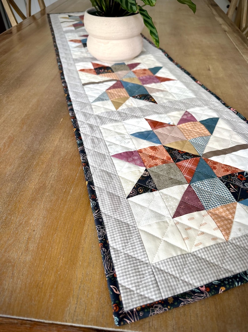 May include: A rectangular quilted table runner featuring a repeating star pattern made of various fabric patches in shades of blue, orange, red, and cream. The runner has a gray and white checkered border and a dark floral print edge.