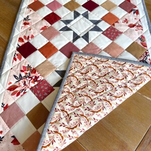 May include: A quilted table runner with a starburst design in the center. The runner features a patchwork pattern of squares in shades of white, red, pink, and brown. The reverse side has a floral pattern. The runner is approximately 12 inches wide.