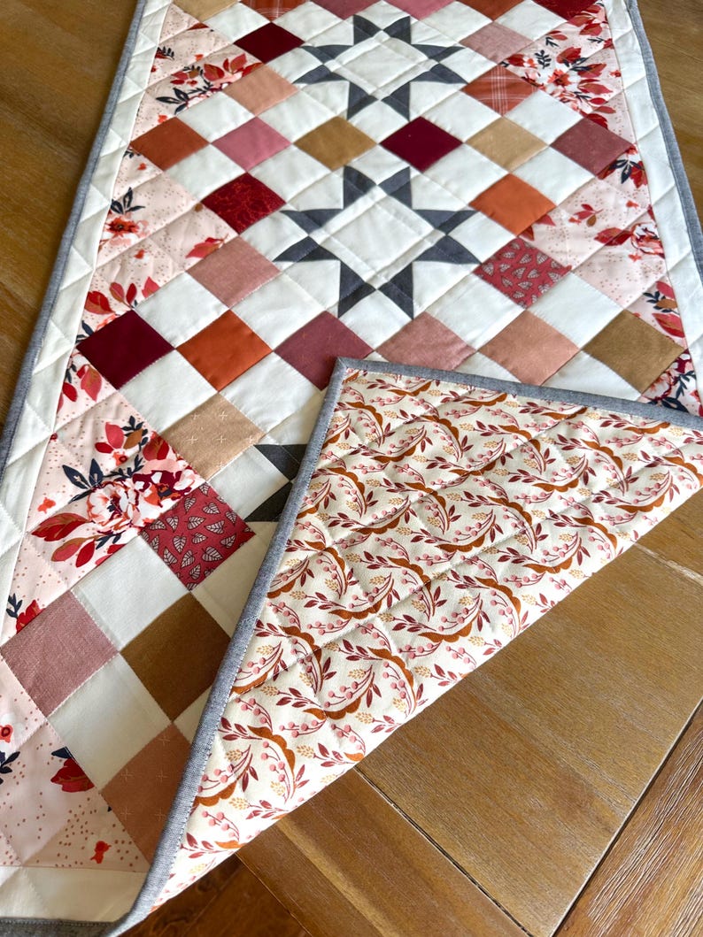 May include: A quilted table runner with a starburst design in the center. The runner features a patchwork pattern of squares in shades of white, red, pink, and brown. The reverse side has a floral pattern. The runner is approximately 12 inches wide.