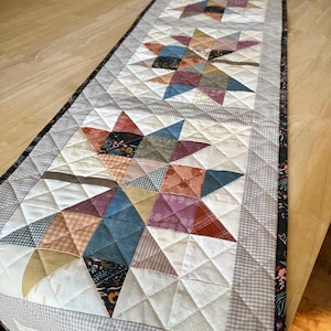 May include: A rectangular quilted table runner featuring three repeating star patterns. The stars are constructed from various fabric squares in shades of blue, red, yellow, and brown, set against a white background with a gray gingham border.