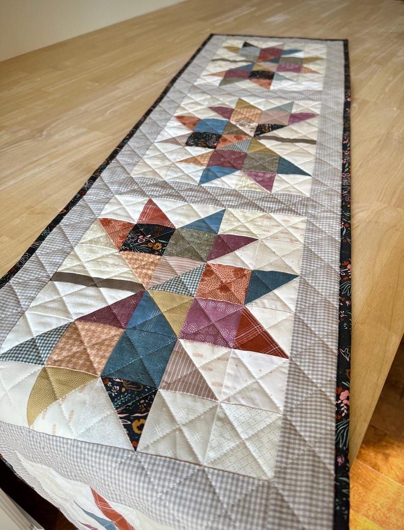 May include: A rectangular quilted table runner featuring three repeating star patterns. The stars are constructed from various fabric squares in shades of blue, red, yellow, and brown, set against a white background with a gray gingham border.