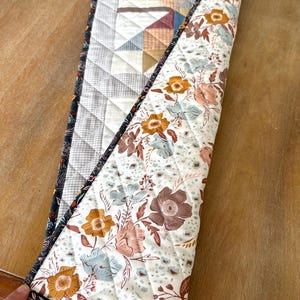 May include: A quilted table runner featuring a star pattern on one side with light gray and muted earth tones, and a floral print on the reverse side with white, orange, and blue flowers. The runner has a dark floral border and diamond stitching.