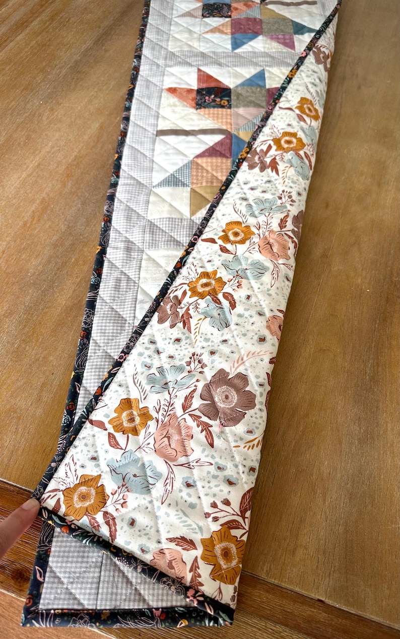 May include: A quilted table runner featuring a star pattern on one side with light gray and muted earth tones, and a floral print on the reverse side with white, orange, and blue flowers. The runner has a dark floral border and diamond stitching.