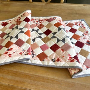 May include: Three quilted table runners with a patchwork design. The runners feature squares in shades of red, pink, orange, and tan, with floral accents and a gray border. The design includes star patterns.