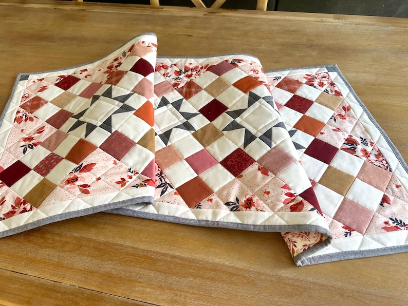 May include: Three quilted table runners with a patchwork design. The runners feature squares in shades of red, pink, orange, and tan, with floral accents and a gray border. The design includes star patterns.