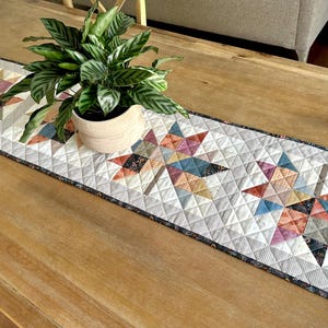 May include: A rectangular quilted table runner featuring a repeating pattern of three large maple leaf motifs. The leaves are constructed from various fabric patches in shades of blue, orange, pink, and grey, set against a white background.