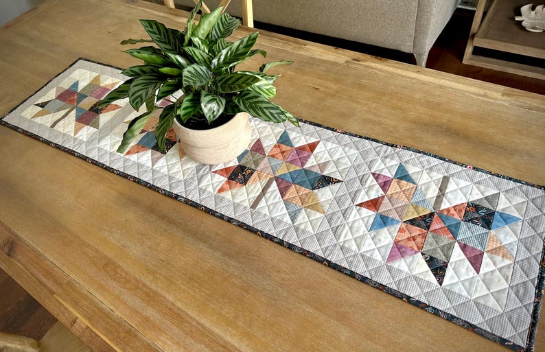 May include: A rectangular quilted table runner featuring a repeating pattern of three large maple leaf motifs. The leaves are constructed from various fabric patches in shades of blue, orange, pink, and grey, set against a white background.