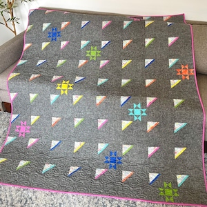 May include: A gray quilt with a pink border and a patchwork design featuring colorful stars. The quilt is made with a variety of fabrics and has a quilted texture.