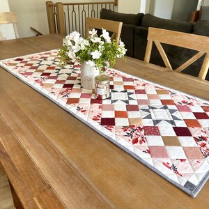 May include: A quilted table runner with a patchwork design in shades of red, pink, and white, accented with floral patterns and a gray border. A vase of white flowers and a candle sit on the runner, displayed on a wooden table.