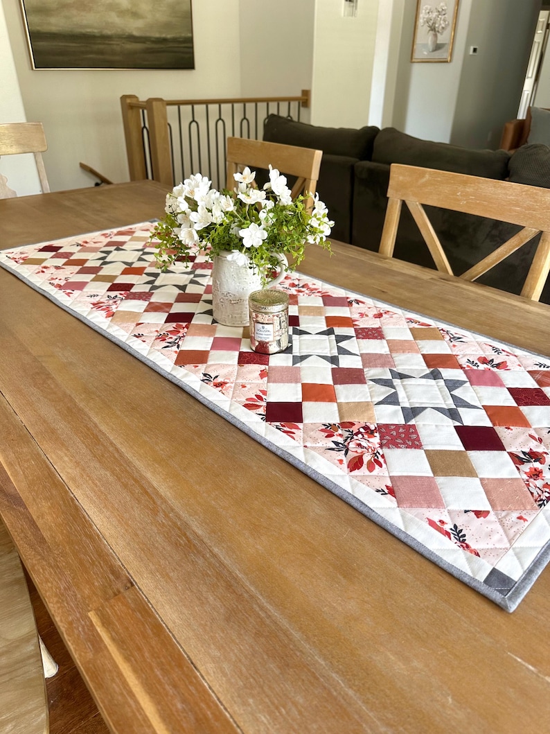 May include: A quilted table runner with a patchwork design in shades of red, pink, and white, accented with floral patterns and a gray border. A vase of white flowers and a candle sit on the runner, displayed on a wooden table.