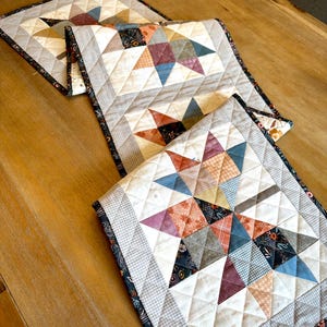 May include: A quilted table runner featuring a repeating star pattern made from various fabric squares in shades of blue, orange, brown, and cream. The runner has a diamond-stitched texture and is finished with a dark floral patterned border.