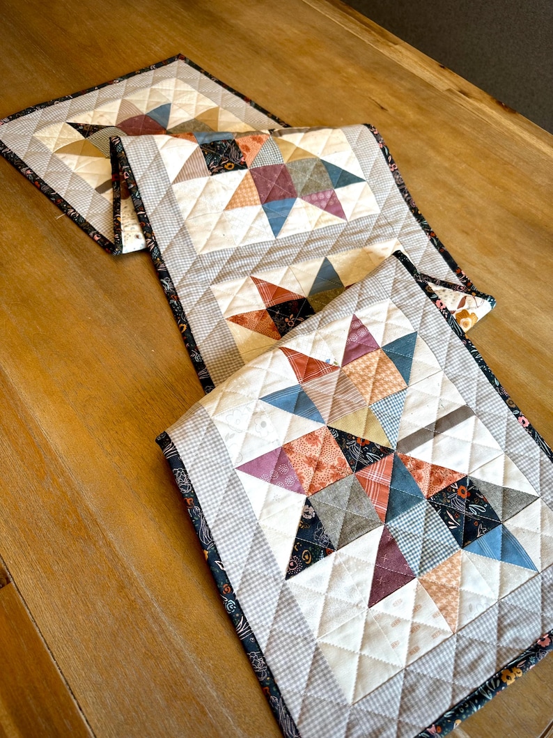 May include: A quilted table runner featuring a repeating star pattern made from various fabric squares in shades of blue, orange, brown, and cream. The runner has a diamond-stitched texture and is finished with a dark floral patterned border.
