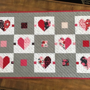 May include: A gray and white quilt with a red border. The quilt is made up of squares with hearts in the center. The hearts are made of different fabrics with pink, red, and black patterns.
