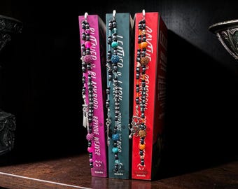 May include: Three books with decorative beaded bookmarks. The books are in shades of pink, teal, and red. The bookmarks feature beads in various colors, charms, and silver-toned metal accents. The books are on a dark wooden shelf.