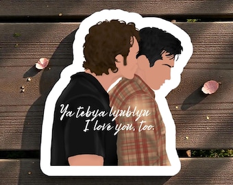 Op de afbeelding: Een sticker met een illustratie van twee mensen die elkaar omarmen. Eén persoon draagt een zwart shirt, de ander een rood-bruin geruit shirt. De tekst "Ya tebya lyublyu I love you, too." staat op de sticker.