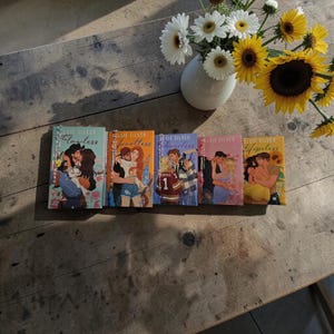 Könnte beinhalten: Fünf Bücher von Elsie Silver, jedes mit einer anderen Coverillustration von Paaren, liegen auf einem Holztisch. Im Hintergrund steht eine Vase mit weißen Gänseblümchen und Sonnenblumen.