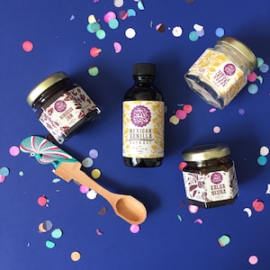 May include: A flat lay of a collection of gourmet food items on a blue background. The items include a jar of hibiscus jam, a bottle of Mexican vanilla extract, a jar of salsa negra, and a jar of vanilla sugar. The items are surrounded by colorful confetti.