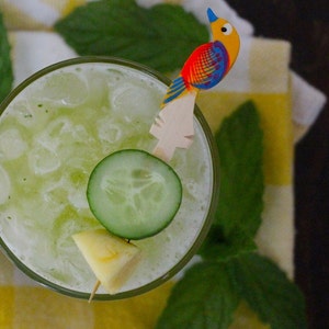 May include: A close-up of a refreshing green drink with ice, cucumber slices, and a pineapple wedge. The drink is garnished with a wooden bird-shaped stirrer.