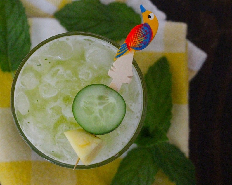May include: A close-up of a refreshing green drink with ice, cucumber slices, and a pineapple wedge. The drink is garnished with a wooden bird-shaped stirrer.