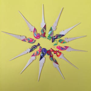 May include: A set of 12 colorful wooden bird toothpick holders. Each toothpick holder features a different bird design with bright colors and intricate details. The toothpicks are arranged in a starburst pattern on a yellow background.