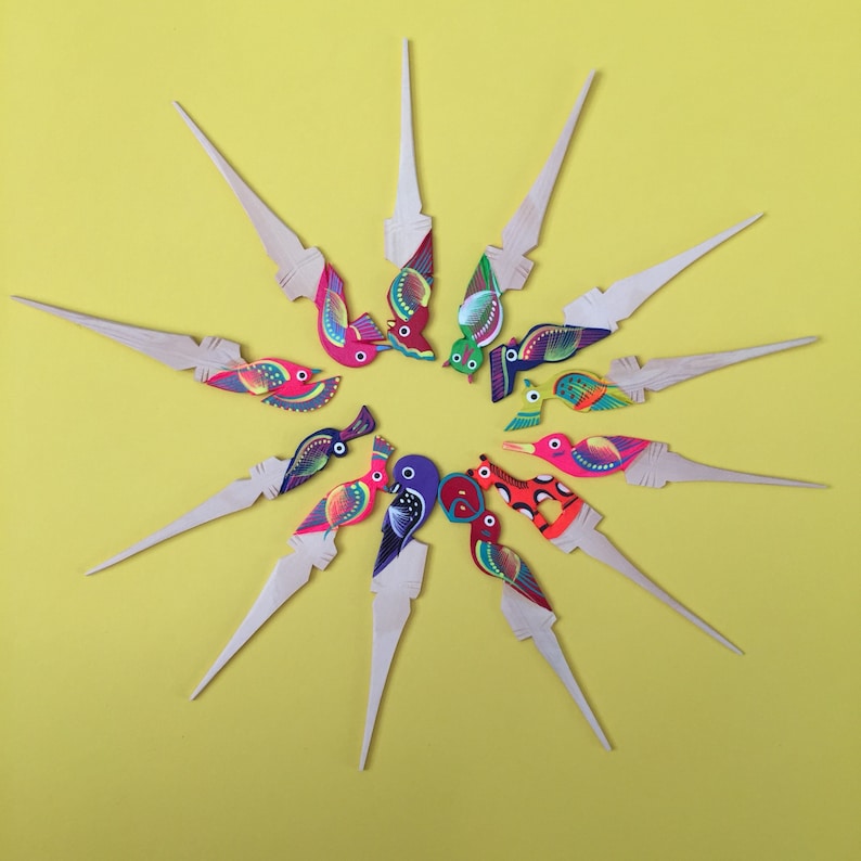 May include: A set of 12 colorful wooden bird toothpick holders. Each toothpick holder features a different bird design with bright colors and intricate details. The toothpicks are arranged in a starburst pattern on a yellow background.