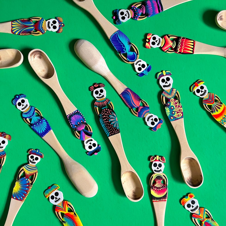 May include: A collection of wooden spoons decorated with colourful Day of the Dead sugar skull designs. The spoons are painted with intricate details and vibrant colours, including pink, blue, green, yellow, and purple.