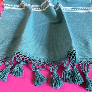May include: Teal blue table runner with white embroidered details and a fringe trim with tassels. The runner is made of a soft, woven fabric.