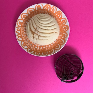 May include: A white concha bread roll on a decorative orange and white plate. A metal concha mold is on a pink surface.