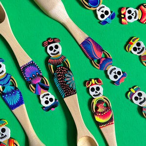 May include: Wooden spoons decorated with colourful Day of the Dead skull designs. The spoons feature a variety of skull designs with intricate details and bright colours, including pink, blue, green, yellow, and black.