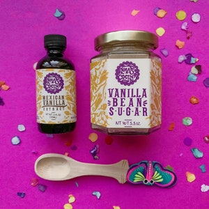 May include: A set of two glass jars with gold lids, containing vanilla bean sugar and Mexican vanilla extract. The jars have colorful labels with the text "CLAS COCINA" and the product names. A wooden spoon with a butterfly design is also included.