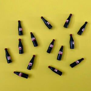 May include: Miniature black bottles with red caps and white labels featuring the Coca-Cola logo are scattered on a yellow background. The bottles are arranged at various angles, creating a playful and eye-catching display.