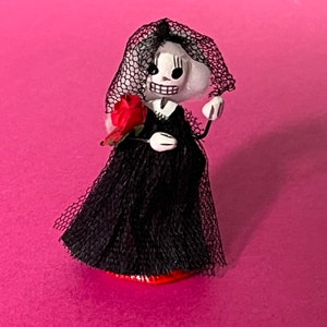 May include: A black and white skeleton bride doll wearing a black tulle dress and veil, holding a red rose.