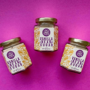 May include: Three glass jars with gold lids and white labels. The labels have a purple logo that says "Las Cocina" and the text "Vanilla Bean Sugar" with a net weight of 5.3 oz.