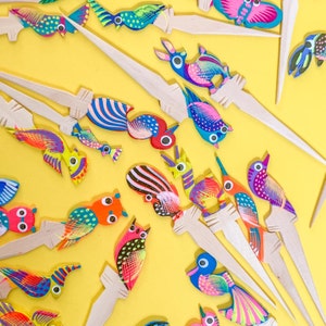 May include: A collection of colorful wooden bird ornaments with intricate details. Each bird is painted with bright colors and patterns, and they are attached to wooden sticks. The birds are arranged on a yellow background.