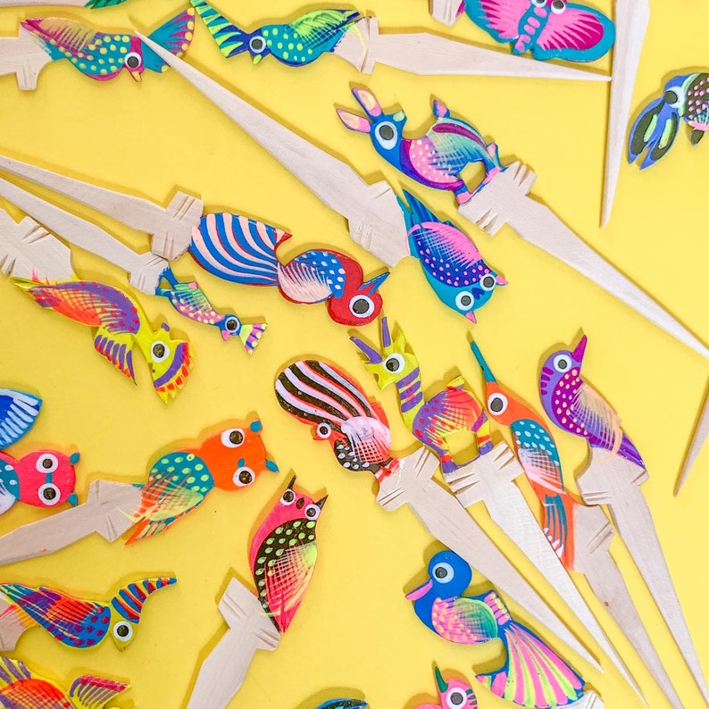 May include: A collection of colorful wooden bird ornaments with intricate details. Each bird is painted with bright colors and patterns, and they are attached to wooden sticks. The birds are arranged on a yellow background.