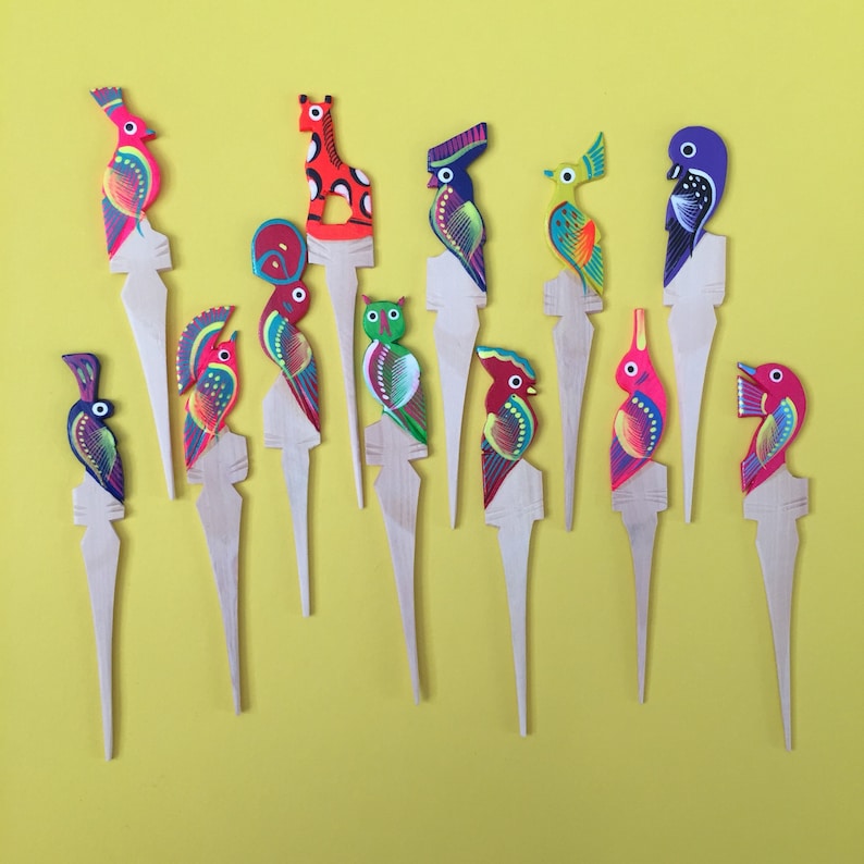 May include: A set of 12 colorful wooden bird and animal toothpick holders. The holders are painted in bright colors, including red, yellow, blue, green, and purple. They are all on a yellow background.