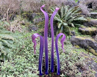 Könnte beinhalten: Vier hohe, schlanke Glasskulpturen in Violett- und Weißtönen, die in einer Gartenlandschaft stehen. Die Skulpturen sind aus Glas gefertigt und haben eine fließende, organische Form.