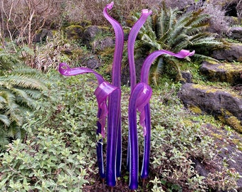 May include: A group of five tall, purple glass sculptures with a swirling, organic design. The sculptures are arranged in a cluster and are set against a backdrop of green foliage and rocks.
