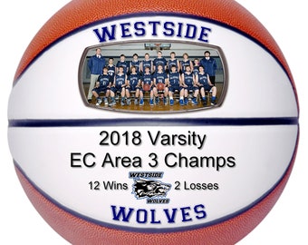 May include: A white basketball with a blue and orange design. The basketball has a photo of a basketball team in the center, with the text "WESTSIDE 2018 Varsity EC Area 3 Champs 12 Wins 2 Losses WOLVES" printed on it.