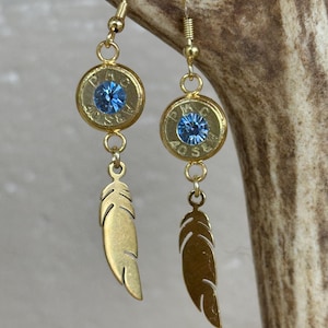 Può includere: Un paio di orecchini color oro. Ogni orecchino presenta un bossolo dorato con un centro di pietra blu, sospeso a un gancio dorato. Sotto il bossolo pende un ciondolo a forma di piuma dorata. Gli orecchini sono esposti su uno sfondo neutro.