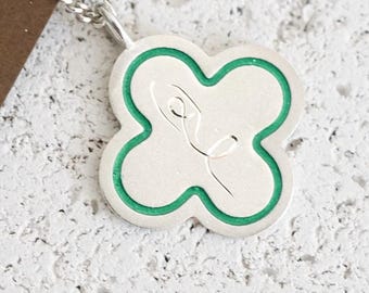 May include: Silver-toned clover pendant necklace with a green border and a minimalist design. The pendant hangs from a delicate chain, set against a textured white and brown background. The necklace is a piece of jewelry.
