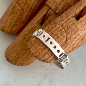 Puede incluir: Un anillo de plata con una serie de cortes rectangulares y circulares. El anillo se muestra en un modelo de mano de madera sobre una superficie de mármol blanco. Diseño sencillo y moderno.