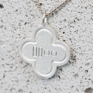 May include: A silver necklace featuring a four-leaf clover pendant. The pendant has a textured finish and the letters "IIloo" are engraved in the centre. The chain is fine and appears to be made of silver.