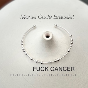 Pode incluir: Uma pulseira de código Morse prateada com a mensagem "FUCK CANCER" escrita em contas. A pulseira é aberta e repousa sobre uma superfície branca. As palavras "Morse Code Bracelet" estão acima da pulseira.