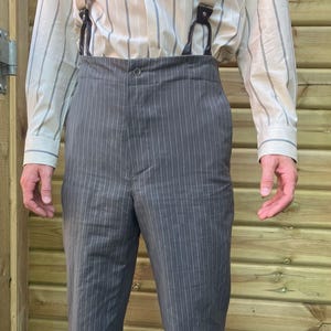 Puede incluir: Pantalones grises de raya diplomática con cintura alta, combinados con una camisa abotonada a rayas color crema y tirantes. Los pantalones tienen cierre de botón y un diseño clásico. Los tirantes son de cuero marrón.