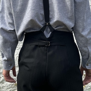May include: High-waisted black trousers featuring a subtle pinstripe pattern, a rear cinch buckle strap, and a V-shaped back yoke. The trousers are paired with a light gray long-sleeved button-down shirt and a single black suspender strap.