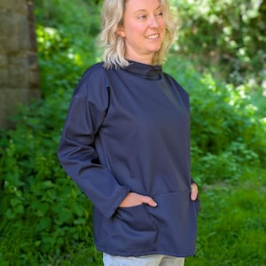 May include: A navy blue long-sleeved top with a high neckline and two front pockets. The top is made of a soft material and is worn with light-wash jeans. The background features green foliage and a stone structure.