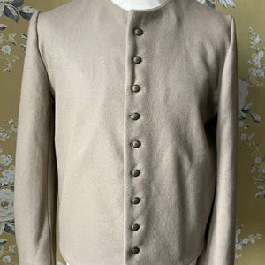 Könnte beinhalten: Beigefarbene Jacke mit rundem Kragen und langen Ärmeln. Die Jacke hat sieben goldfarbene Knöpfe. Das Kleidungsstück wird auf einer Schaufensterpuppe präsentiert. Der Hintergrund hat ein Blumenmuster.