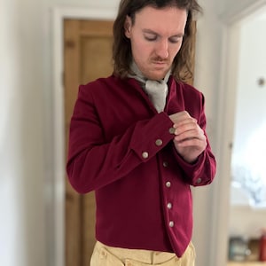 Könnte beinhalten: Eine weinrote Wolljacke mit Stehkragen und einer Reihe von Zierknöpfen. Die Jacke hat eine figurbetonte Silhouette und lange Ärmel.